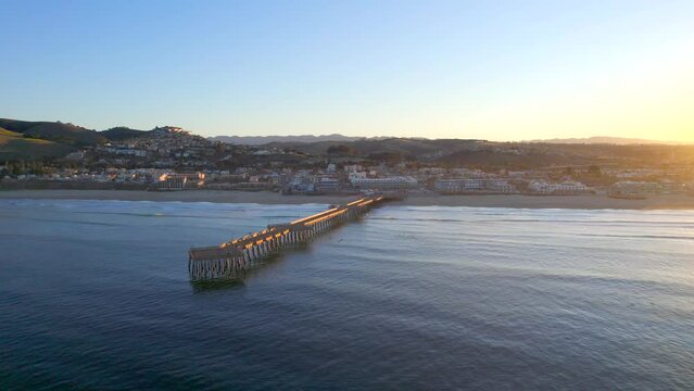 Aerial view of Pismo Beach, California with a pier jutting out from the beach into the water. Drone shot flying slowly towards the shore at sunrise.