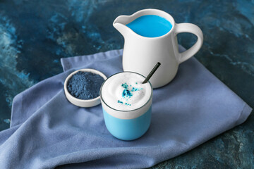 Glass and jug of blue matcha tea and bowl with powder on blue grunge background