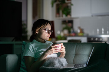 Focused young woman in glasses using laptop sitting on couch at home in the night. Thoughtful girl...
