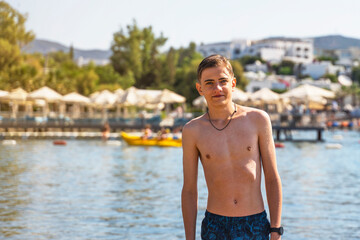 Young handsome teenager in swimsuit walking at the beach with blue sky and sea water in background.