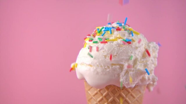 wafer cone with scoop of ice cream covering and strewing sprinkles on pink background, copy space, slow motion, close up