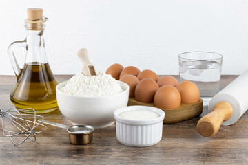 Baking ingredients: butter, flour, sugar, cold water, eggs, baking powder, whisk, rolling pin. Ingredients for making a cake or cupcake on a wooden background, side view.