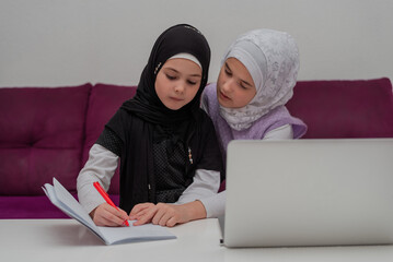 Two child girls in the hijab together learn for school at the home desk. Sister helping little sister to do homework. Young Muslim students have online education.