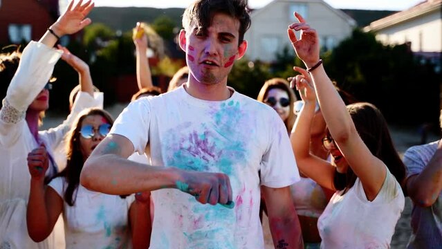 Handsome man stained in colored powder punches cheast, shows peace sign with his fingers at Holi festival. Girls dance, strike guy down to ground, laugh. End of lockdown, covid pandemic, restrictions