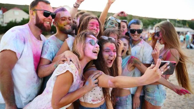 Cheerful people covered in colorful powder take selfie on smartphone and smile at Holi festival on beach in slow motion. Friends smeared in dry colors take group photo on phone. End of covid pandemic