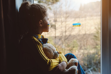 Sad little girl kid with flag of ukraine on face holding fluffy bear sitting alone on windowsill at home. Protest against russian war invasion in Ukraine. Ukrainian Flag, Protesters concept