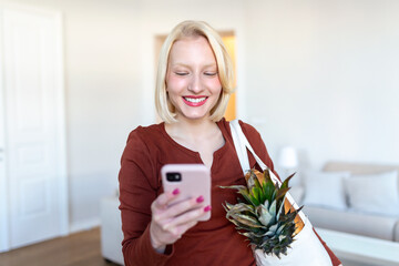 Pretty blond woman arriving home carrying groceries in a eco friendly tote bag using her smart mobile phone - Multi tasking