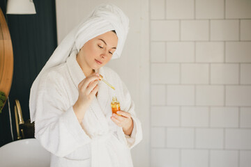 Young woman wearing white bathrobe and towel on head holding face oil in bathroom. Beauty cosmetic...