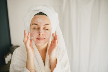 Young beautiful woman wearing bathrobe and towel on her hair applying moistrizing cream on her face. Skin care morning rituals. Beauty routine.