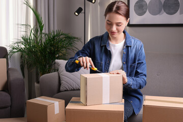 Young woman using utility knife to open parcel at home - obrazy, fototapety, plakaty