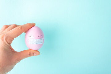 One pink colorful Easter egg with painted eyes and with protective mask covid-19 in the hand. The concept of celebrating Easter on quarantine. Selective focus