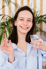 Smiling young woman holding pill omega capsule and glass of water indoors. Girl taking medicine at...