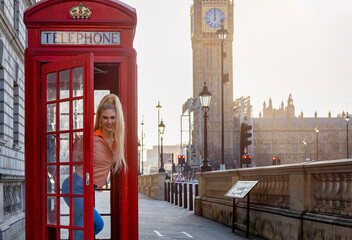 A blonde tourist woman stands in a classic, red telephone booth in front of the Big Ben clocktower...