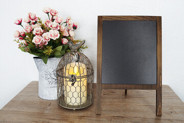 Empty blackboard with bird cage and flower in metal vase on wooden shelves