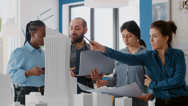 Close up of colleagues using blueprints, laptop and tablet to design building model for project. Group of people doing teamwork to plan construction layout with urban maquette.