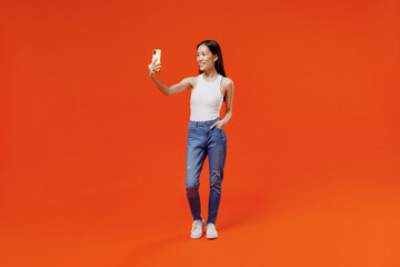 Full size young woman of Asian ethnicity 20s in white tank top get video call use mobile cell phone do selfie talk conducting pleasant conversation isolated on plain orange background studio portrait.