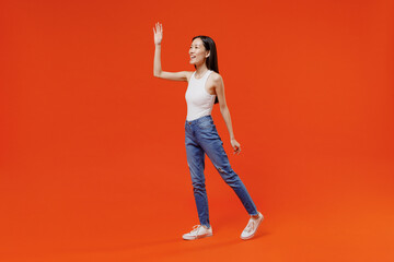 Fototapeta na wymiar Full size body length side view charming young woman of Asian ethnicity 20s years old in white tank top meet greet waving hand as notices someone isolated on plain orange background studio portrait.