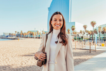 Young happy smiling attractive businesswoman standing staring at camera, at beach of Barcelona city street to office, skyscrapers cityscape and beach with palms background. Copy space. - obrazy, fototapety, plakaty