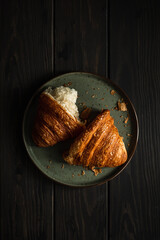 A freshly baked and torn apart delicious croissant on a plate on a wooden table, shot in dark and moody style with natural light, from above, selective focus