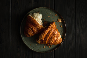A freshly baked and torn apart delicious croissant on a plate on a wooden table, shot in dark and moody style with natural light, from above, selective focus