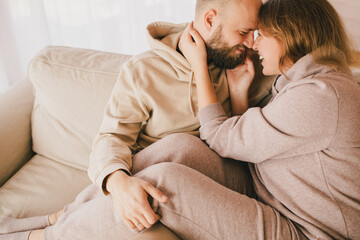 Couple in love wearing beige casual suits sitting on couch in cozy wooden house, hugging and kissing.