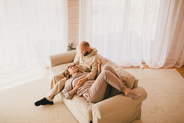 Couple in love wearing beige casual suits sitting on couch in cozy wooden house, hugging and kissing.