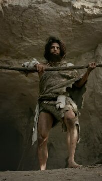 Primeval Caveman Wearing Animal Skin and Fur Hunting with a Stone Tipped Spear in the Prehistoric Cave. Prehistoric Neanderthal Hunter Scavenging. Video Footage with Vertical Screen Orientation
