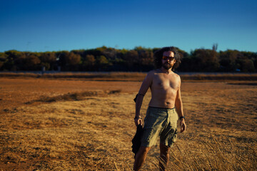 Man with long hair and sunglasses poses shirtless. Adventurer stands in the savannah. Bracelets and...