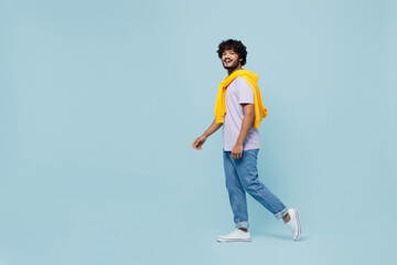 Full size body length side profile view cheerful blithesome young bearded Indian man 20s years old wears white t-shirt stepping walking isolated on plain pastel light blue background studio portrait.