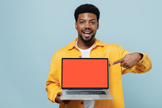 Young happy man of African American ethnicity wear yellow shirt hold use work point index finger on laptop pc computer with blank screen workspace area isolated on plain pastel light blue background.