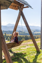 A young woman is sitting and enjoying time, relaxing on a wooden swing in the mountains on a bright summer sunny day