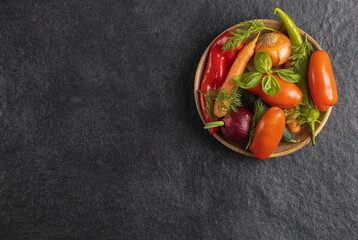 Traditional wooden plate with fresh seasonal vegetables: green pepper, tomato, onion, carrot, cucumber, basil, dill on gray black background, space for text, top view, stock photo