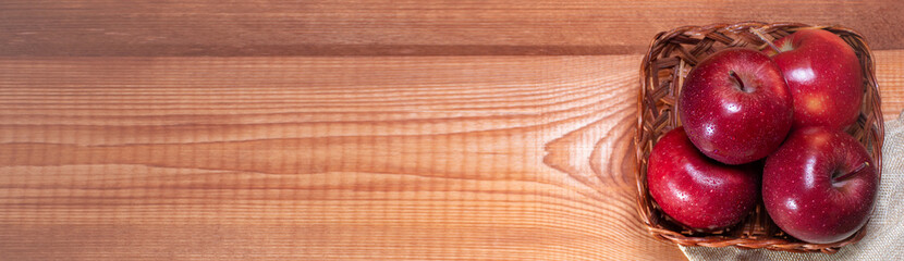 Fresh red apples lie on wooden basket on wooden background next to pink flowers and storage bag with place for text: healthy snack, vitamins, healthy food, ingredients for baking recipes