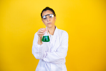 Young brunette woman wearing scientist uniform holding test tube over isolated yellow background relaxed with serious expression on face. simple and natural looking at the camera.