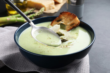 Spoon with delicious asparagus soup over bowl on dark table