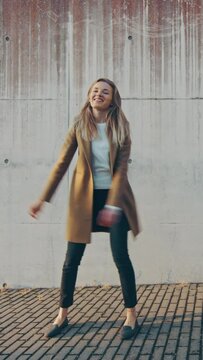 Cheerful and Happy Young Blond Female is Walking and Actively Dancing on a Street next to an Urban Concrete Wall. Vertical Screen Orientation Video