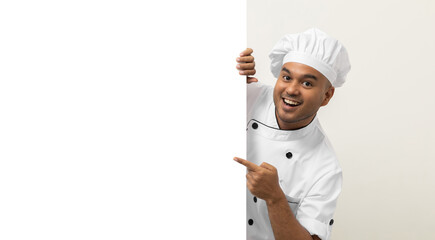 Happy Young handsome asian man chef in uniform looks out from behind an empty whiteboard. Cooking indian man hiding behind big blank billboard for advertising text menu in kitchen and restaurant.