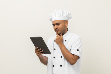 Young asian male chef holding digital tablet thinking to choose various delicious dish menu good taste for customer order. Online delivery food buying variety of dish