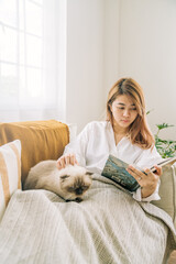 Young girl is rading a book with happy soft cuddly white cat lying in lap enjoying and purring at home warm and cozy atmosphere