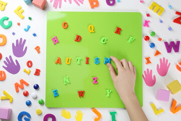 Woman making word Autism on magnetic board at white table, top view