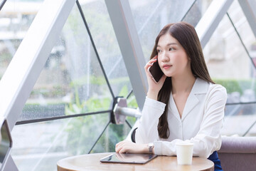 Beautiful young Asian professional business female in a white shirt is  while using your smartphone in your hand to talk she is sitting happy smile working at coffee shop among business background