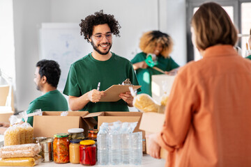 charity, donation and volunteering concept - happy smiling male volunteer with clipboard and woman taking box of food at distribution or refugee assistance center
