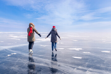 Fototapeta na wymiar Winter lake Baikal Russia, two tourist women friends in red cap are skating on ice frozen, sunny day