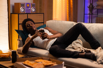 Lazy man lies on the couch relaxed, afro guy muscular sends messages, browses social media, watches...