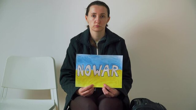 Refugee woman holding motivational packard with text No war against armored conflict on background of Ukrainian flag. Crisis, peace, stop aggression in whole world