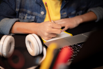 Close-up of girl's hands taking notes with pencil in notebook, laptop, headphones, teenage girl has remote lessons, distance learning, online school