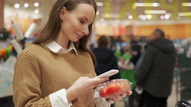 Young woman takes products from the supermarket shelf, holds a smartphone, scans the barcode on the tomato packaging using a mobile phone.