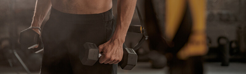 Cropped view of muscular young man holding dumbbells in hands and doing exercise to strengthen his biceps in fitness center