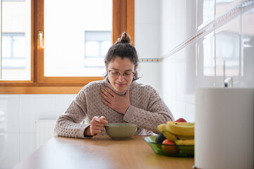 Woman with sore throat trying to eat soup in the kitchen.