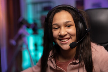 Close-up portrait of a smiling young gamer, the girl is happy to win the game, pass the level, the teenager has a headset, professional gaming accessories, the room lit by fioket blue led lights